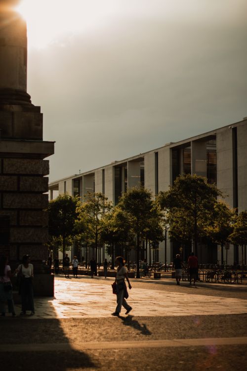 Free Woman walking in a vibrant town square at sunset with shadows from modern architecture. Stock Photo