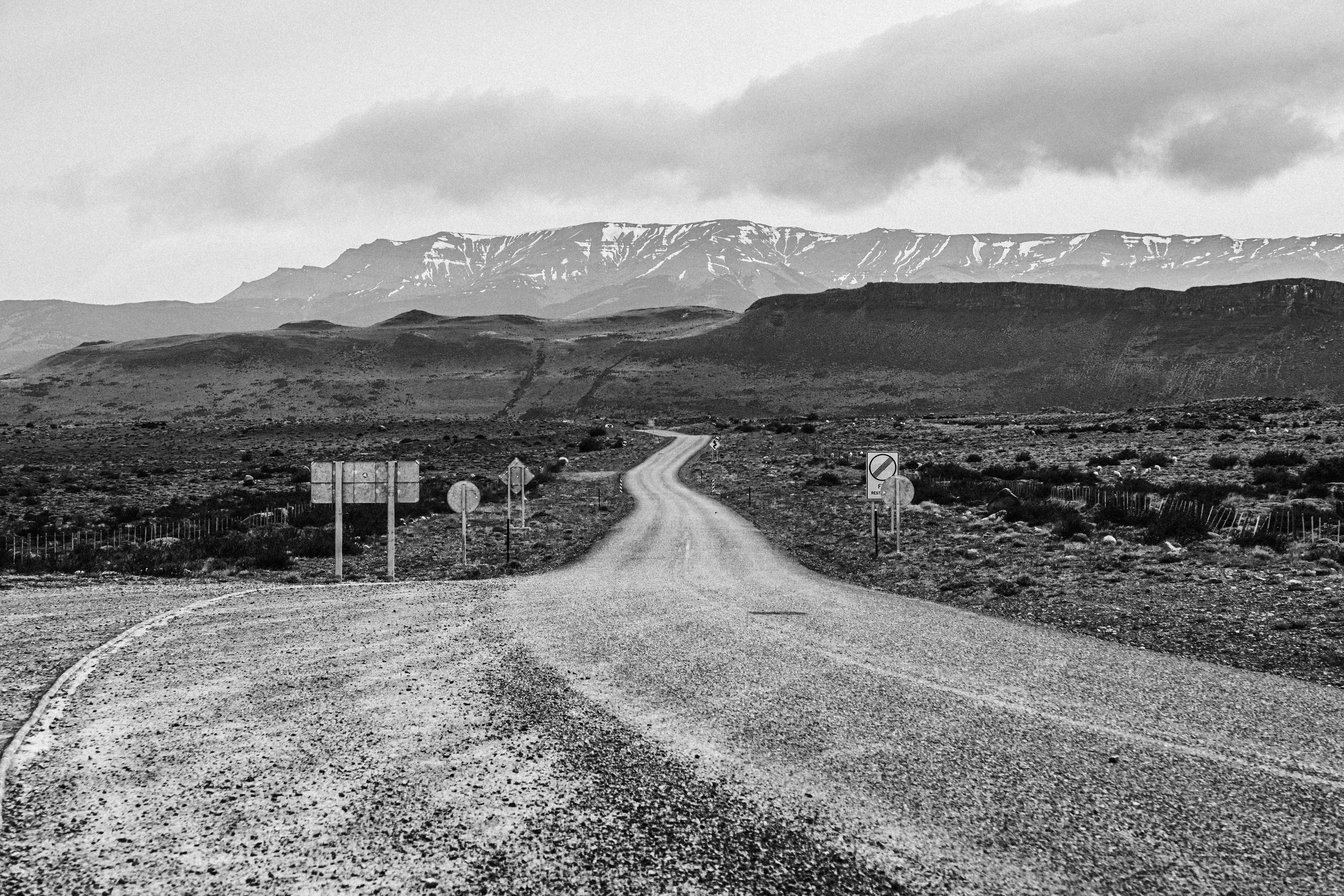 A winding dirt road leading to snow-capped mountains under a moody sky in black and white.