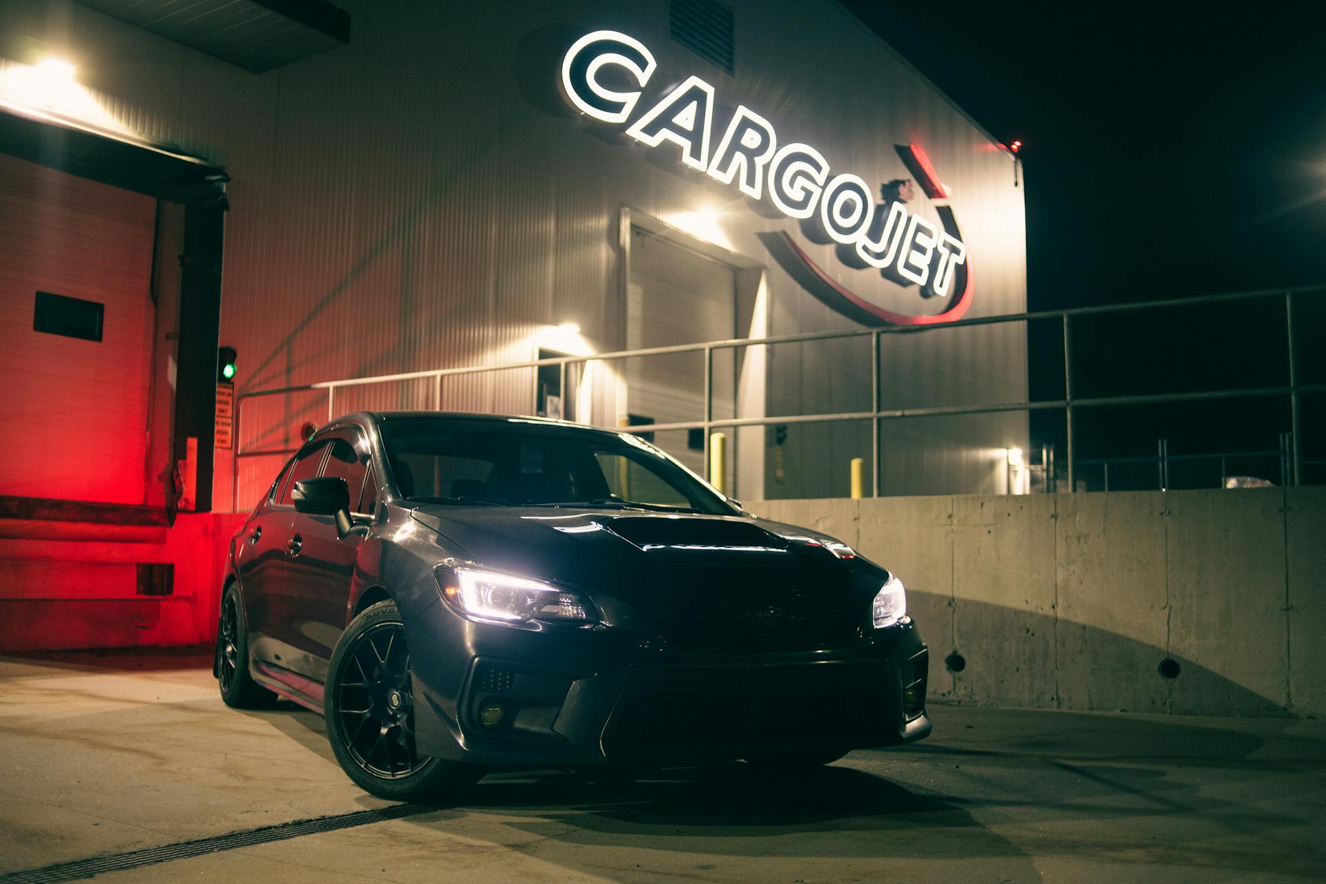 Black car with headlights on, parked at night outside CargoJet facility, under illuminated signage.