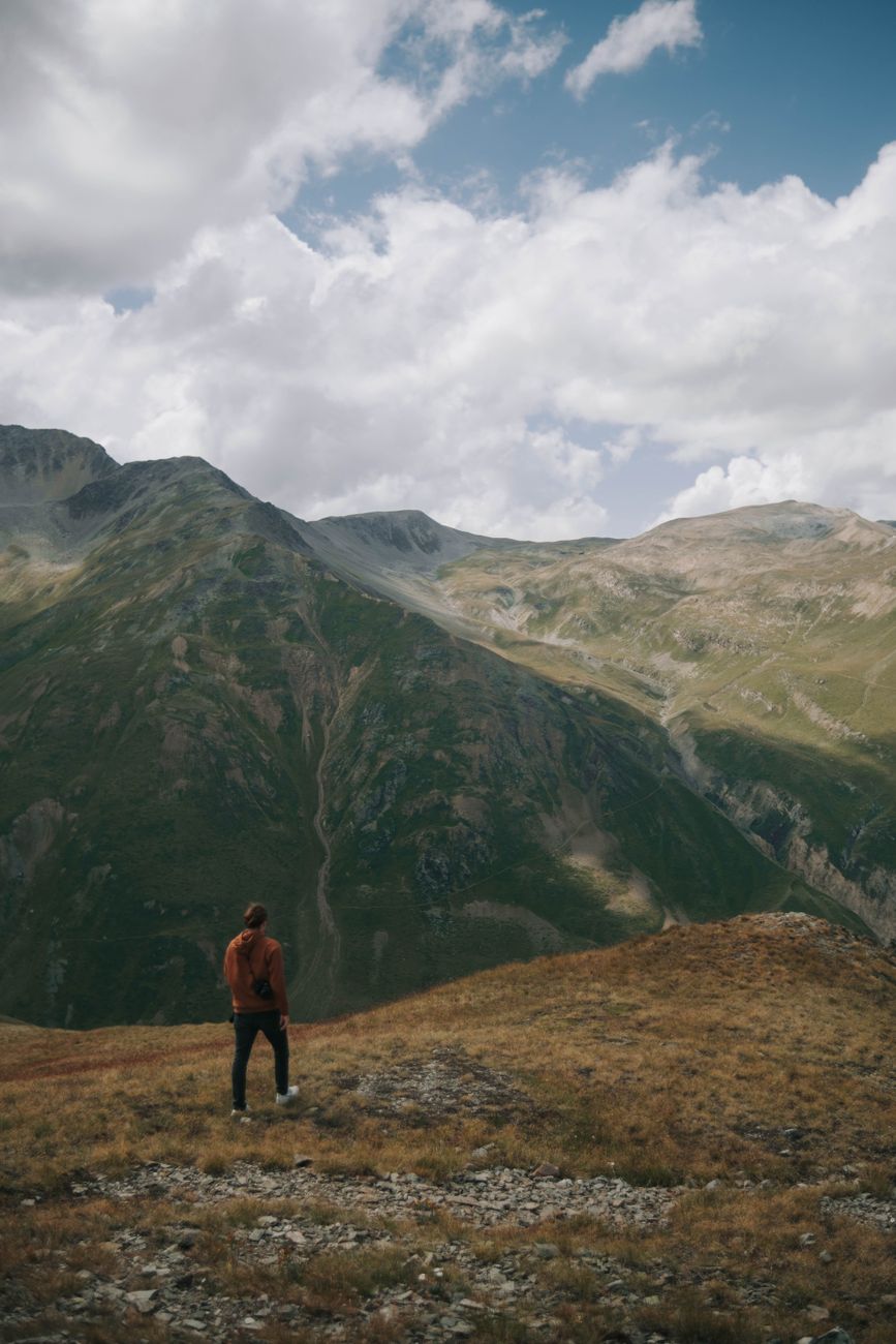 Man standing on a mountain top, gazing at the vast landscape under a cloudy sky.