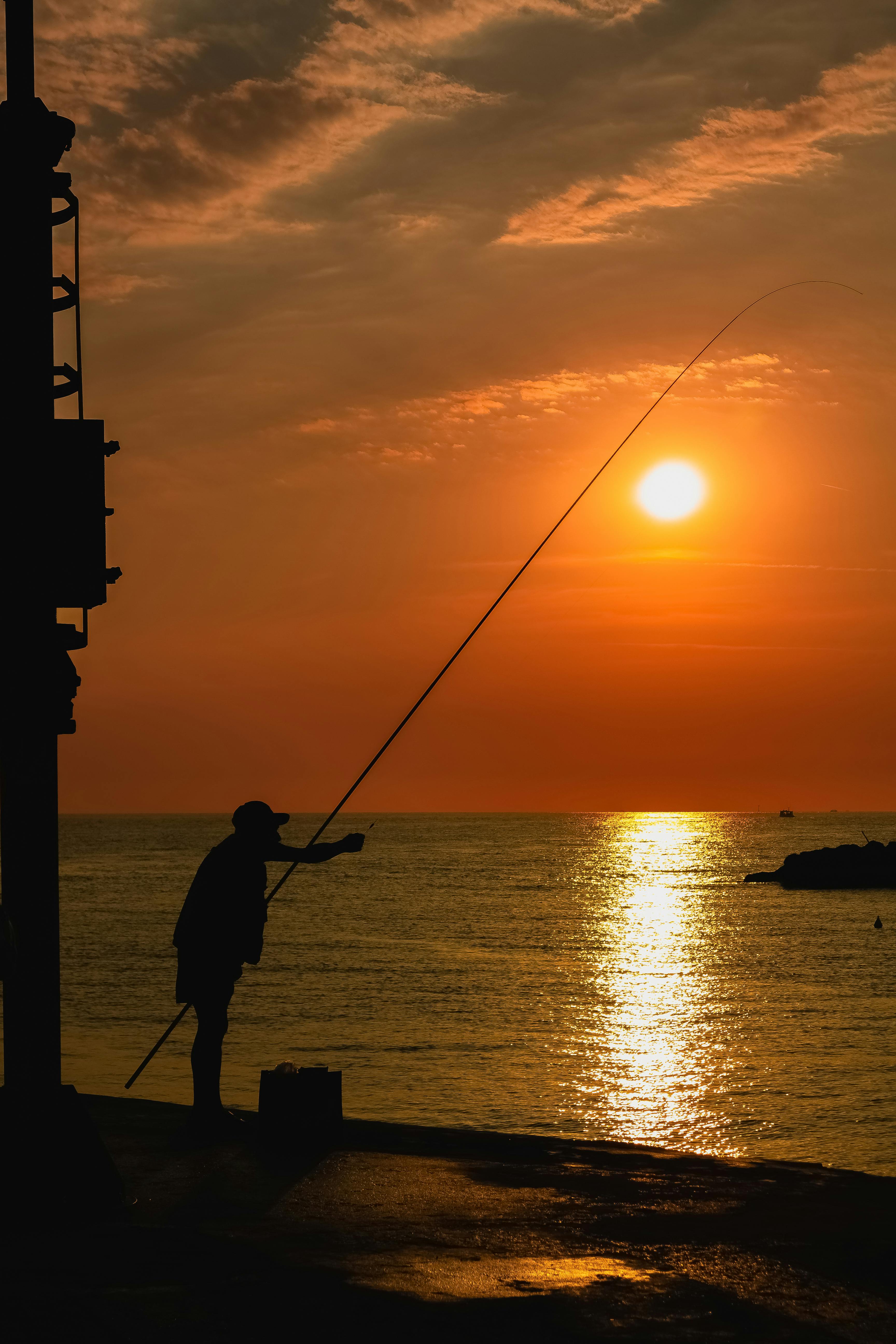 Silhouette Of Man Holding Fishing Rod Standing On A Rock During Sunset ·  Free Stock Photo