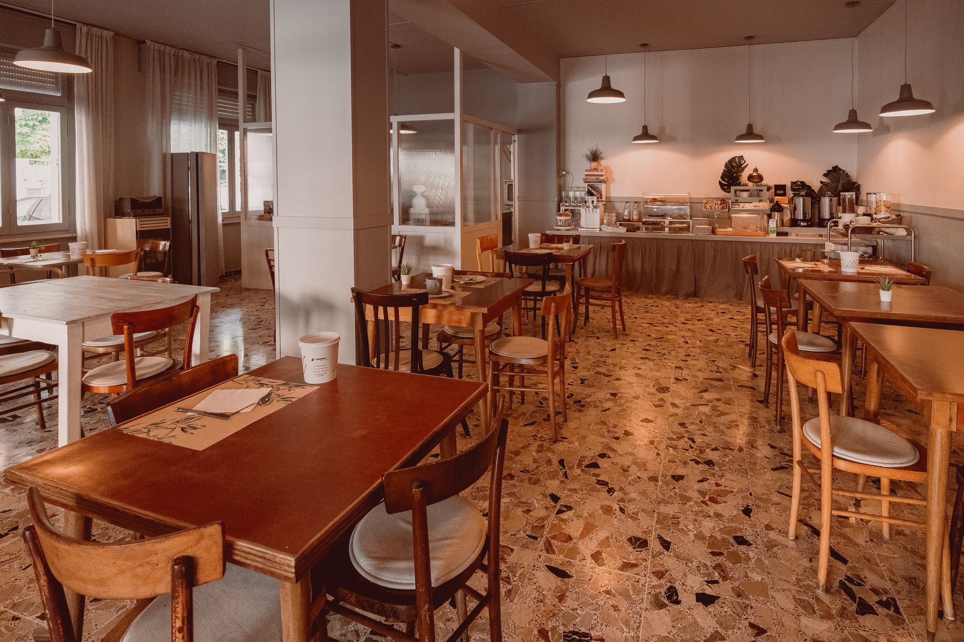 Wide interior of a cozy modern bistro with hanging plants, wooden tables, warm pendant lighting, and large windows.