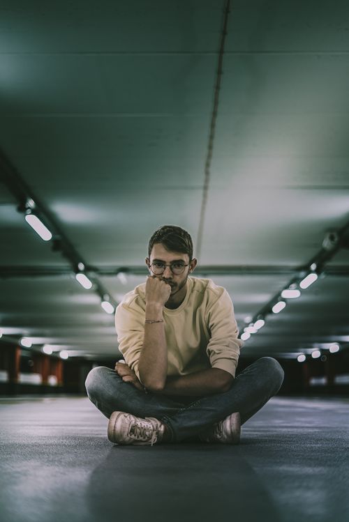 Free A thoughtful man sits cross-legged in a dimly lit urban parking garage offering an introspective scene. Stock Photo