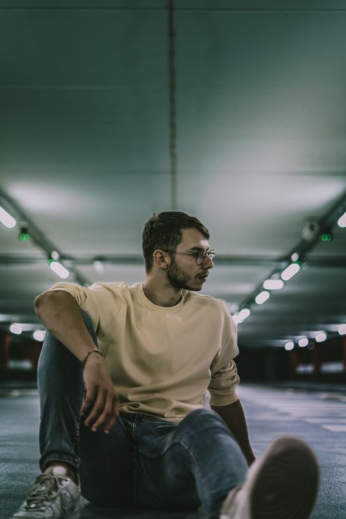 Free A young man sits thoughtfully in a dimly lit urban parking garage. Stock Photo
