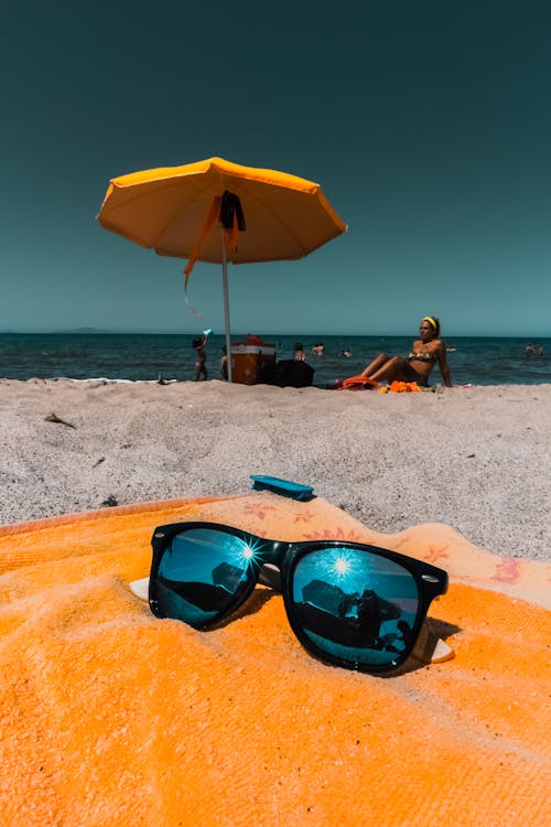 Black Framed Sunglasses on Towel with Woman Sitting on Beach Sand on Background