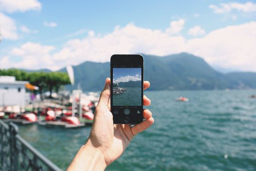 Hand holding smartphone photographing a beautiful lake landscape with mountains.
