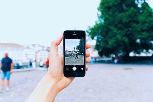 Free A hand holding a smartphone taking a photo of a scenic outdoor setting with people walking by. Stock Photo