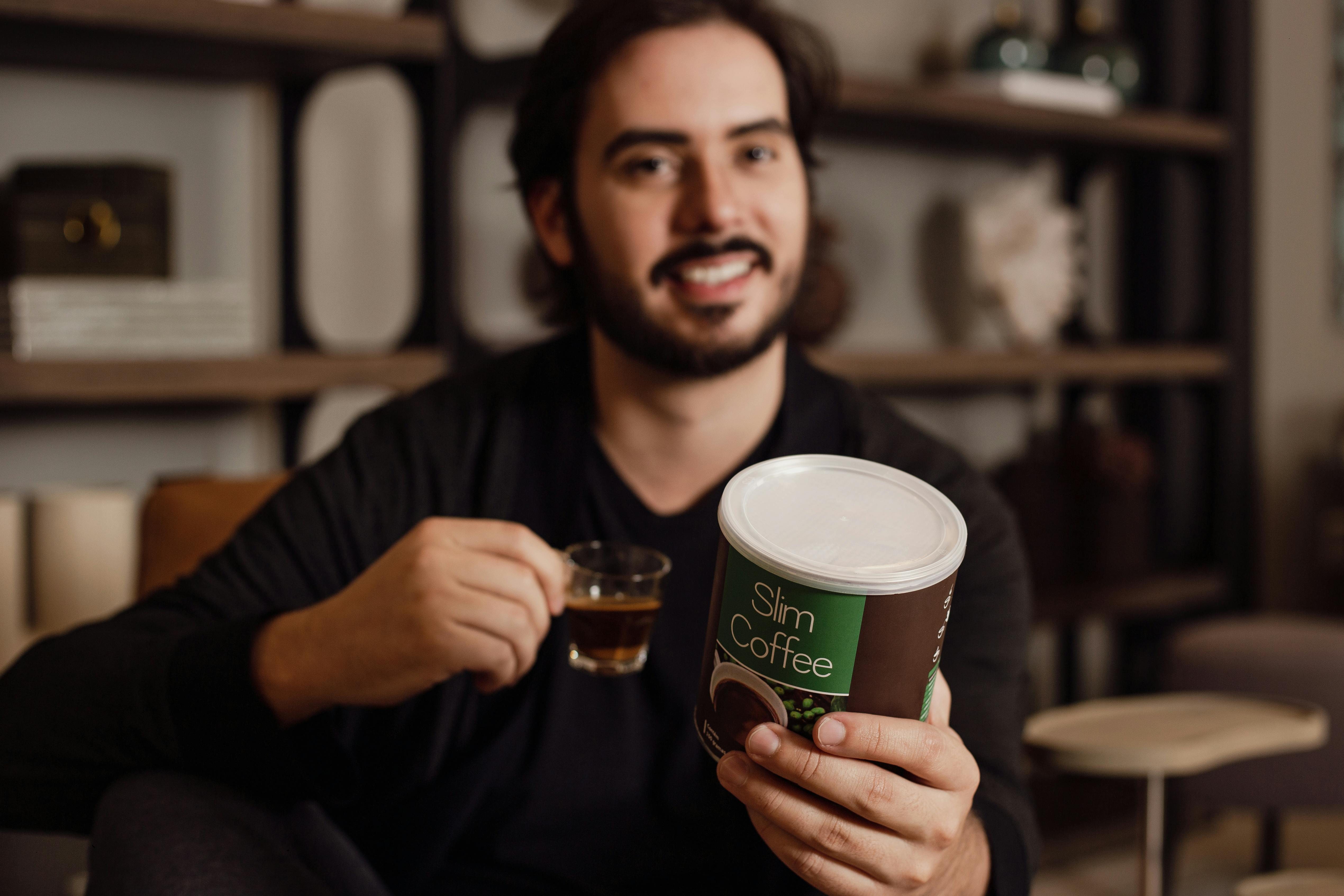 Smiling man holding a Slim Coffee can while enjoying a cup of coffee indoors.