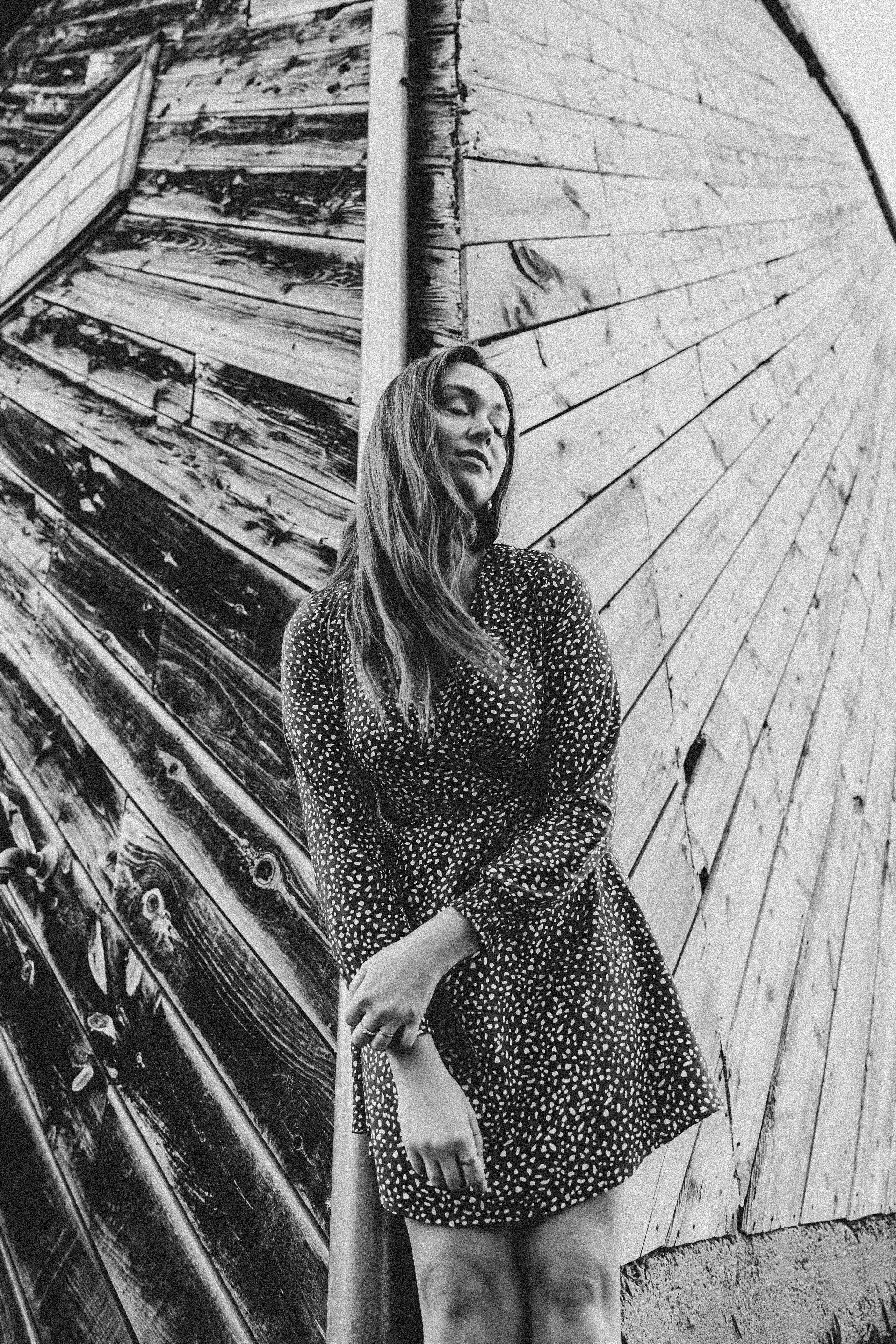 Artistic black and white portrait of a woman standing by a rustic wooden wall.