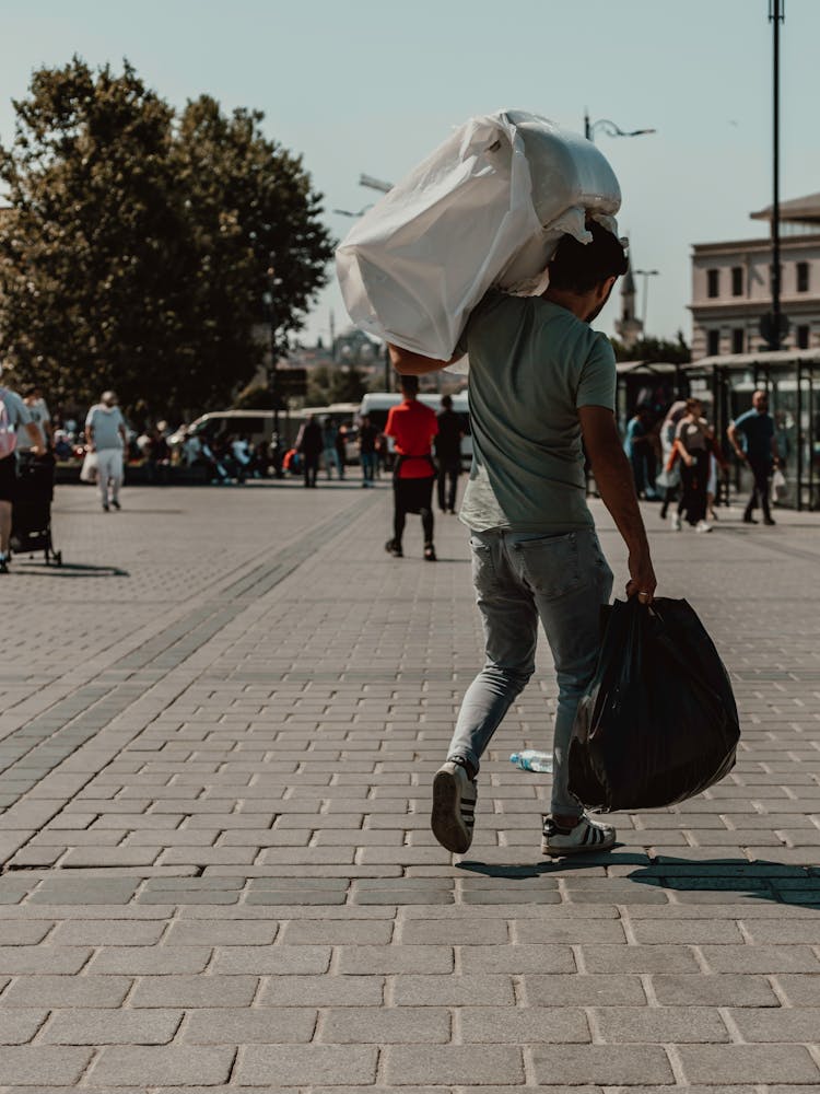 Man In Green T-shirt And Gray Pants Walking On Stone Pavement Carrying Bag And Load On Shoulder