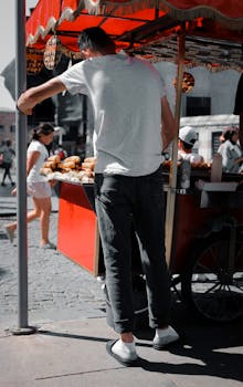 Street vendor selling food at a cart in bustling urban area under a red canopy.
