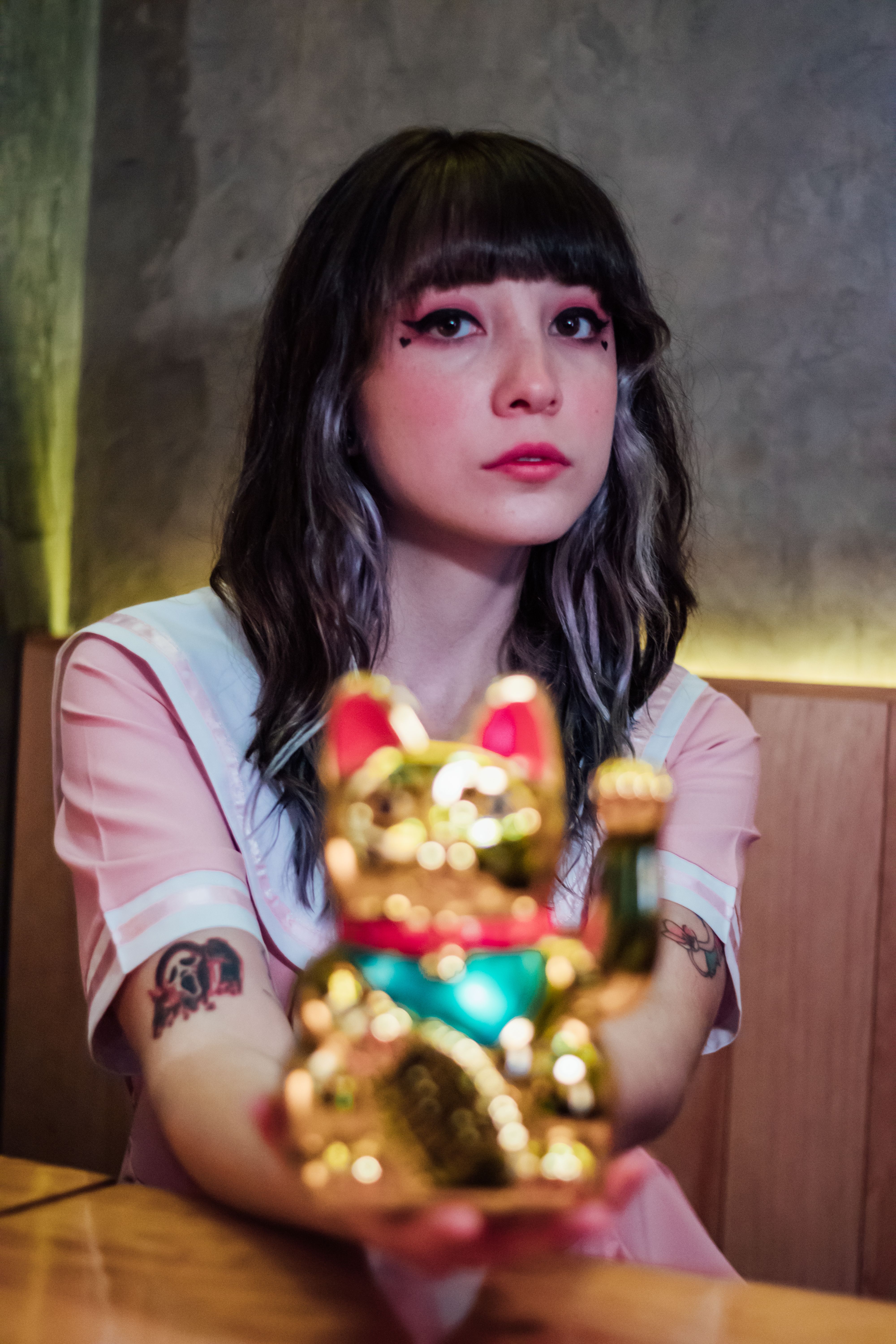 Portrait of a young woman with tattoos holding a golden Maneki-neko figurine indoors.