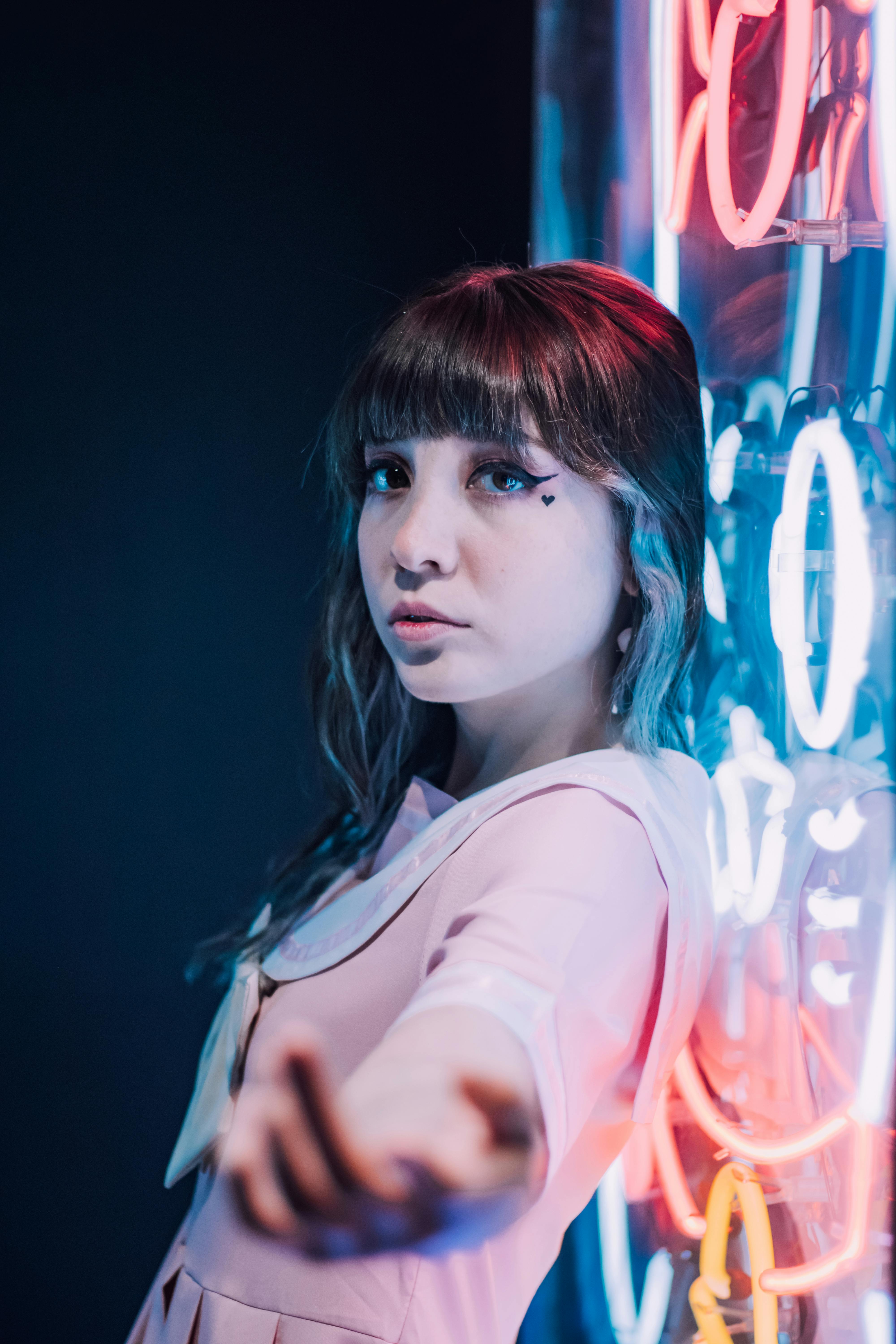 A young woman in a pink dress poses artistically with vibrant neon lights in the background.