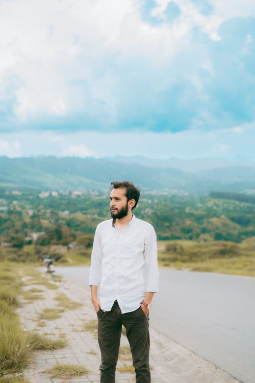 Free Man with a beard in a white shirt standing by a road with scenic green hills in the background. Stock Photo