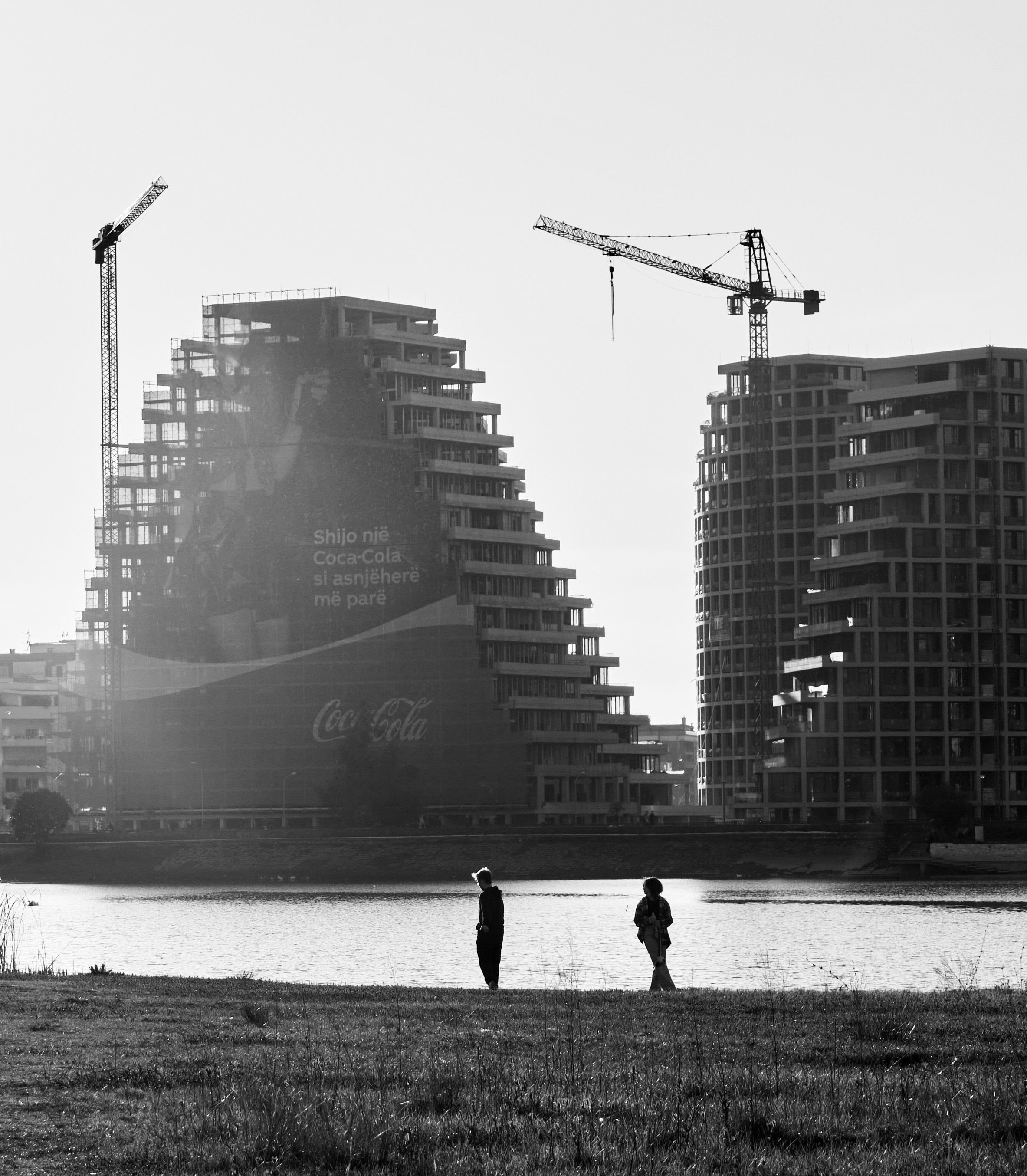 Monochrome view of Tiranë's modern skyline with cranes near a lakeside.