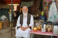 Elderly Afghan vendor selling kettles in Kabul market, sitting outdoors.