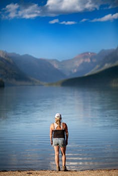 A woman stands by the shore of a tranquil lake with mountains in the background, enjoying a summer day.