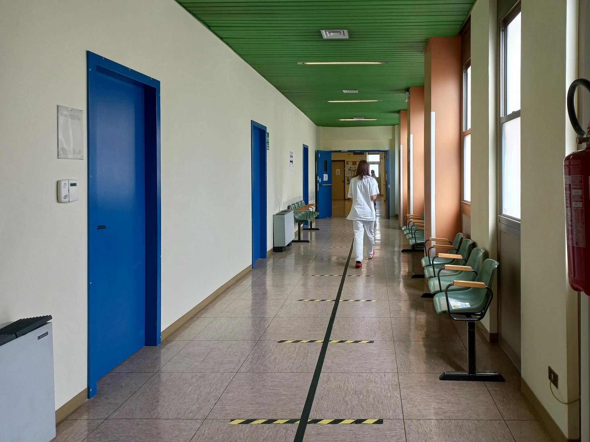 A healthcare worker walking down an empty hospital corridor with blue doors and green ceiling.