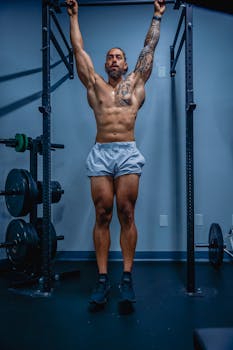 Muscular man with arm tattoos doing pull-ups in a gym, showcasing strength and fitness.