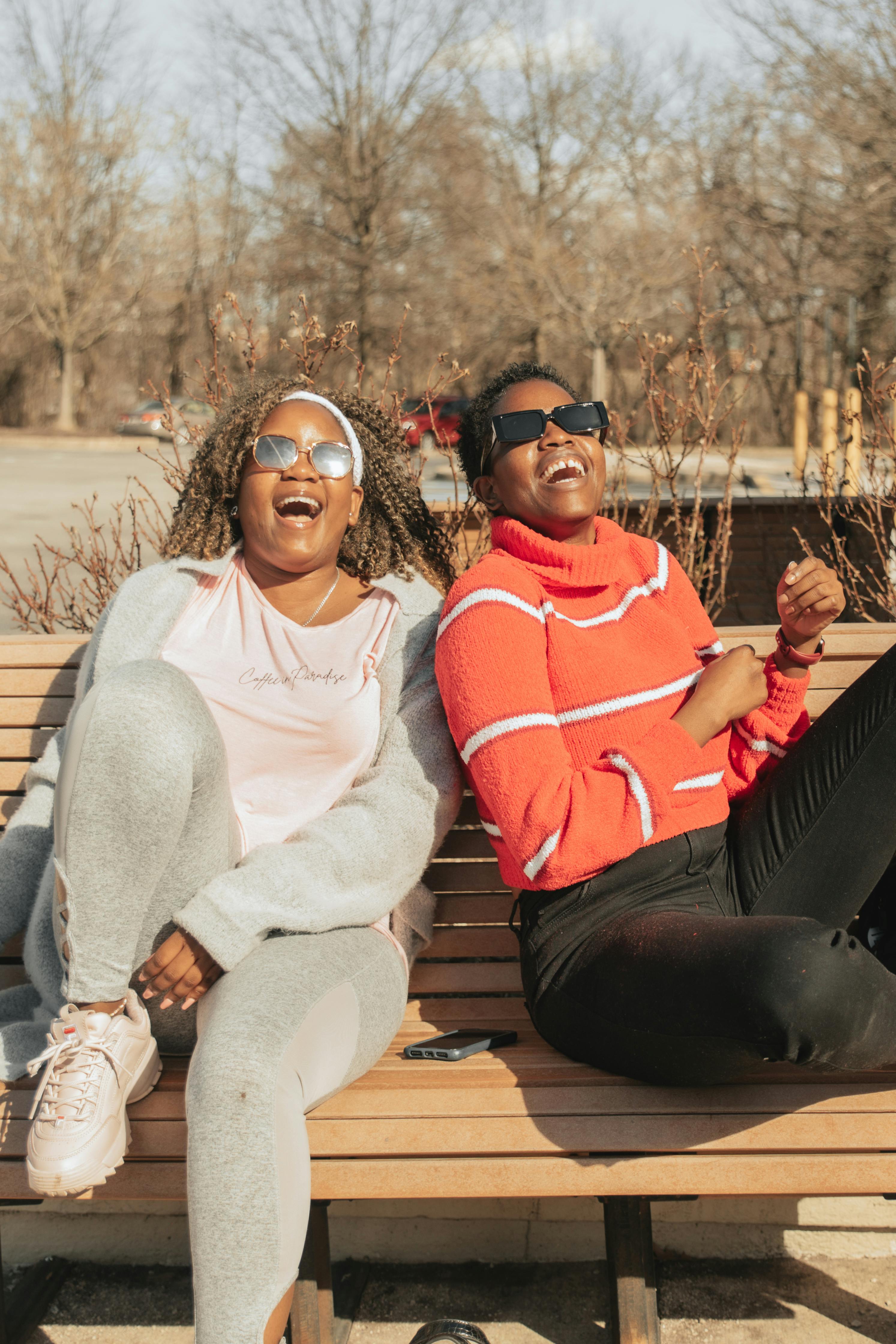 happy women wearing sunglasses laughing while sitting on a wooden bench