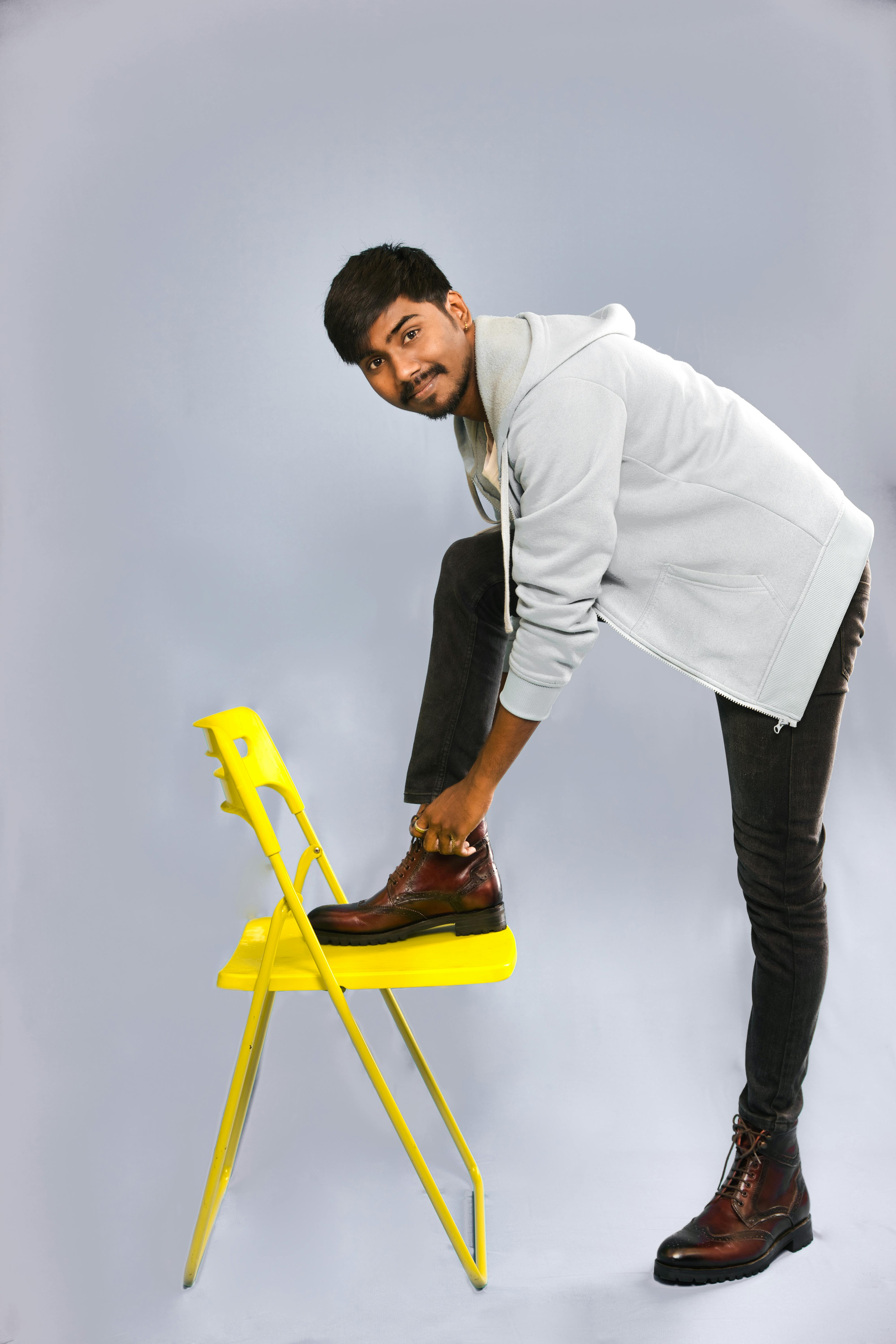 Portrait of a South Asian man in a gray hoodie smiling while posing with a yellow chair in a studio setting.