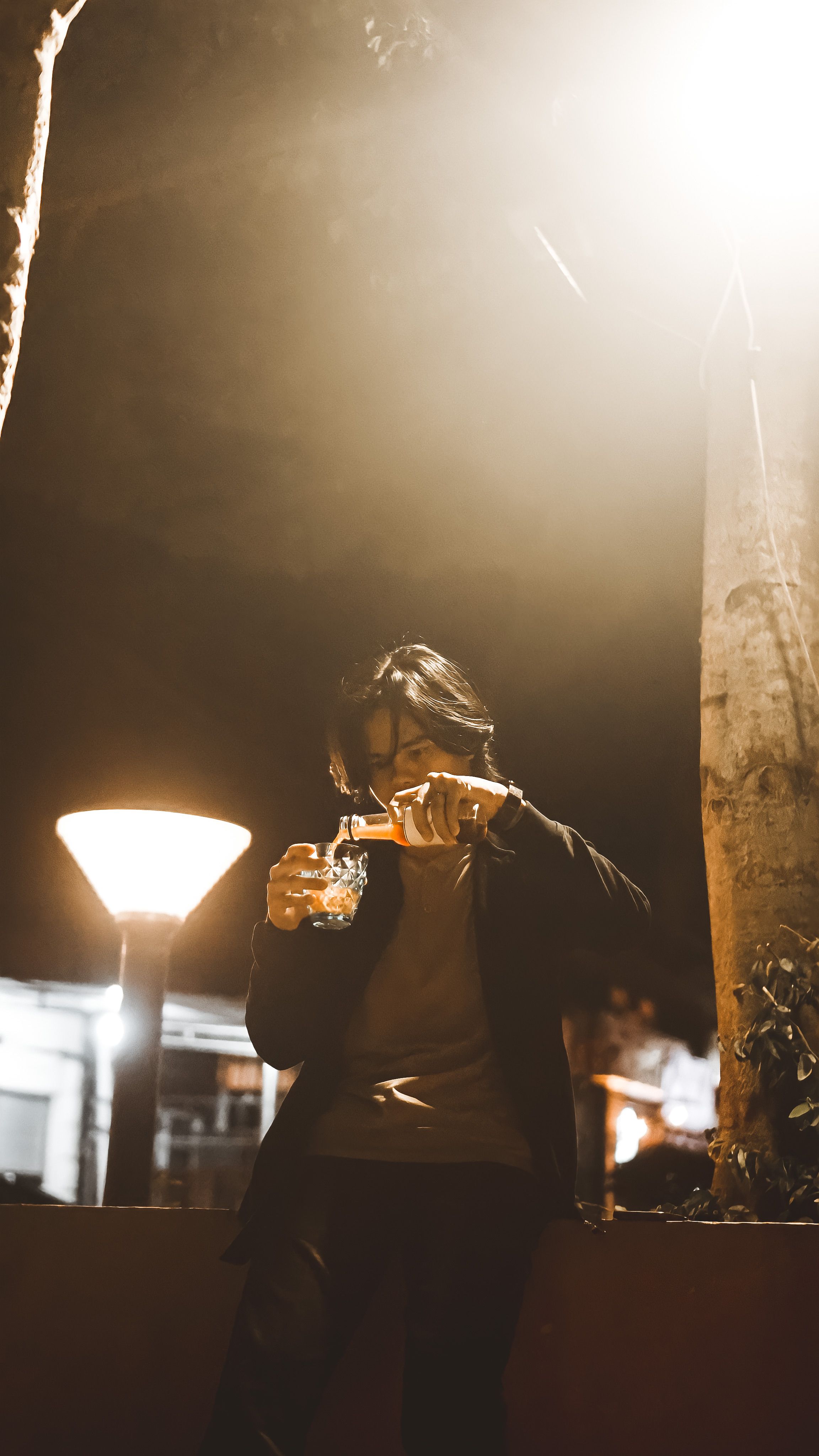 A young man is pouring a beverage from a bottle outdoors under a streetlight at night.