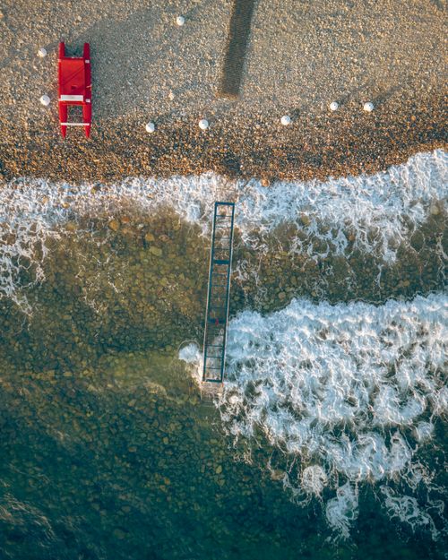 Free Dramatic aerial view of ocean waves hitting a rocky beach, featuring a vibrant red lifeguard seat. Stock Photo