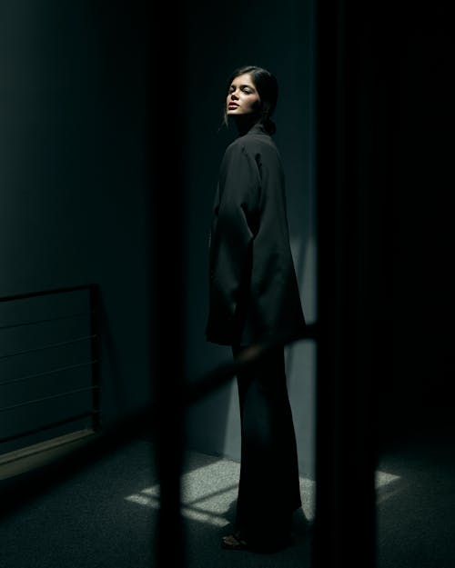Woman in Black Formal Attire Standing in a Dark Room while Looking at the Camera