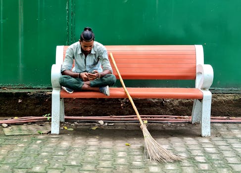 A man sitting on a bench outdoors, using a smartphone with a broom beside him.