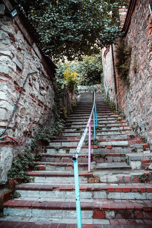 Free A quaint outdoor stairway with brick steps and a colorful metal railing, framed by rustic stone walls and lush greenery. Stock Photo