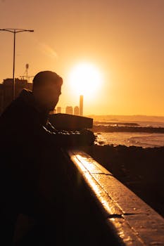 Silhouette of a man leaning on a railing by the sea during sunset, creating a dramatic shadow effect.