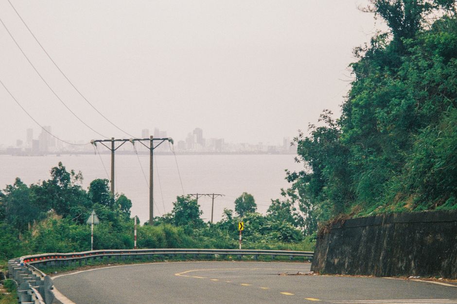 Scenic view of a curvy road with lush greenery, overlooking a distant city skyline and calm sea in Vietnam.