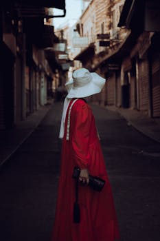 A woman in a red dress and white hat holding a camera stands in an empty street, exuding mystery and elegance.