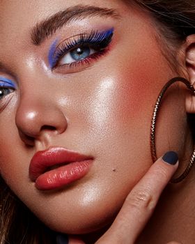 Close-up portrait of a woman with bold makeup and hoop earrings, highlighting vibrant eye shadows and glossy skin.