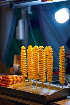 Close-up of spiral fried potatoes on sticks at a lively night market food stall.