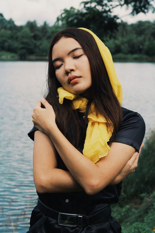 Free A serene portrait of a young woman with closed eyes, wearing a yellow headscarf by a tranquil lake. Stock Photo