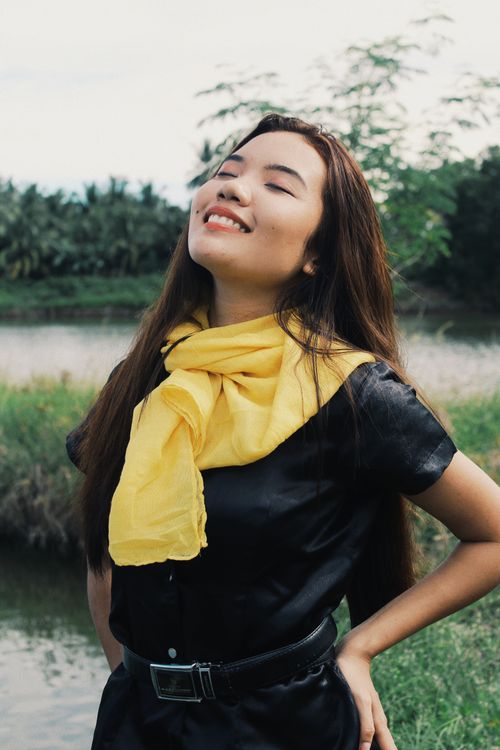 Free Smiling woman with yellow scarf, eyes closed, enjoying nature by a serene lake. Stock Photo