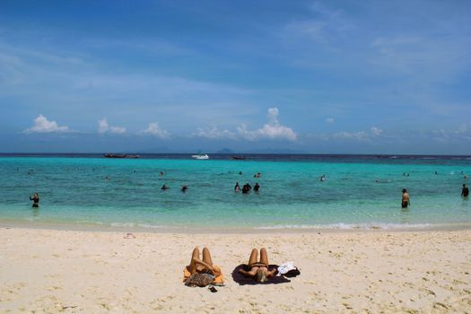 Sunny beach scene in Thailand with people sunbathing and swimming in turquoise waters.