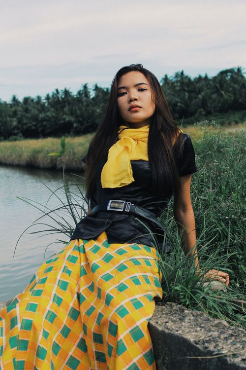 Free A fashionable young woman in a vibrant skirt poses by a serene riverside, showcasing modern style. Stock Photo
