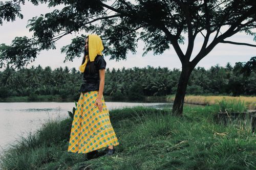 Free A fashionable woman in a vibrant skirt and yellow headscarf stands by a serene lakeside surrounded by lush greenery. Stock Photo