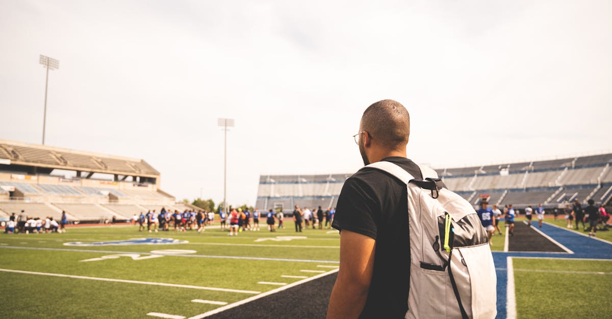 A man with a backpack watches a football practice on a sunny day at a stadium.