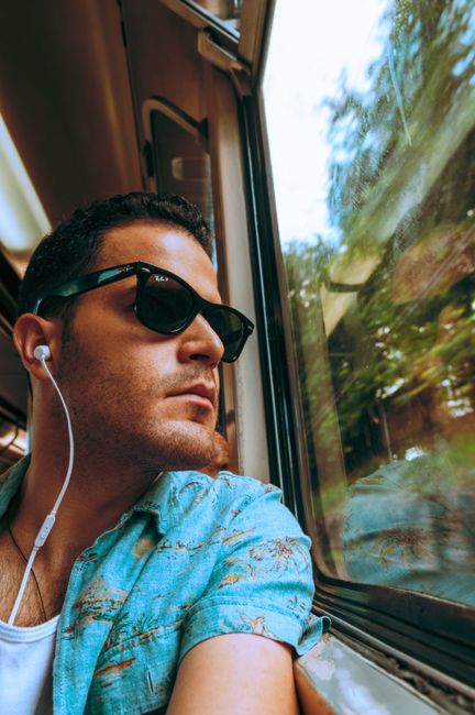 Man with sunglasses listens to music on a train, looking out the window.