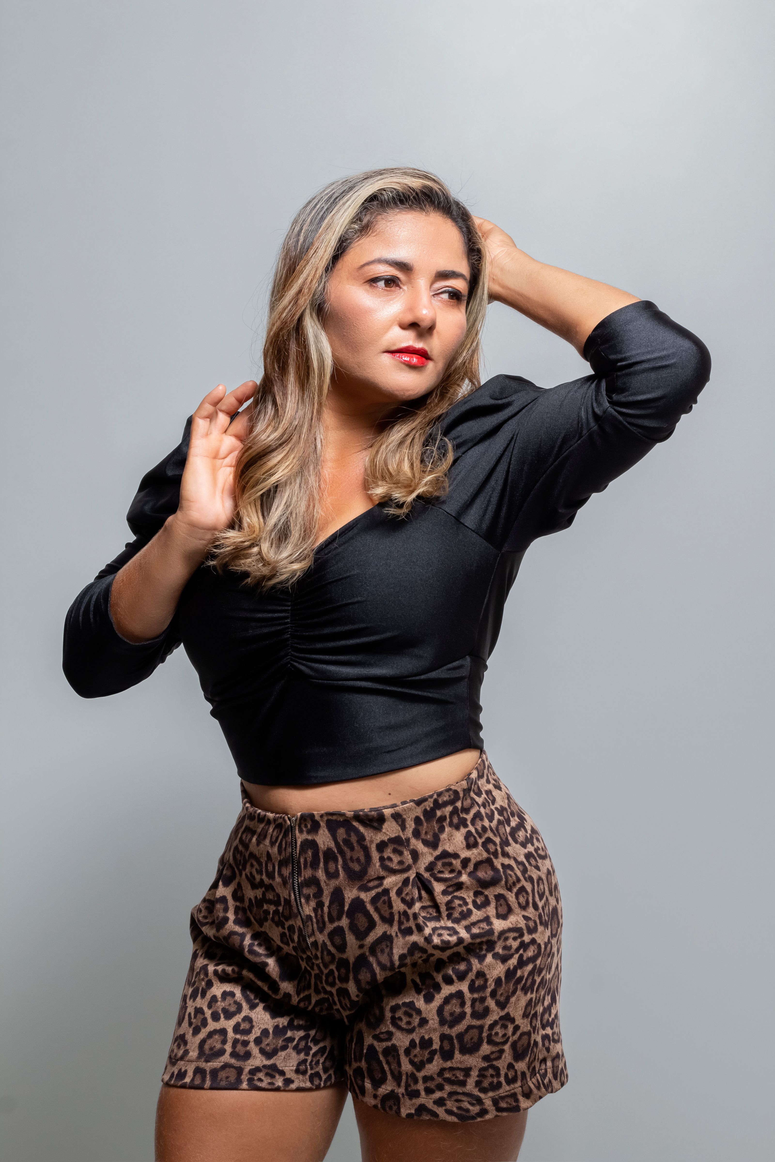 Mulher adulta estilosa com blusa preta e shorts de onça posa em ambiente interno com a mão na cabeça.