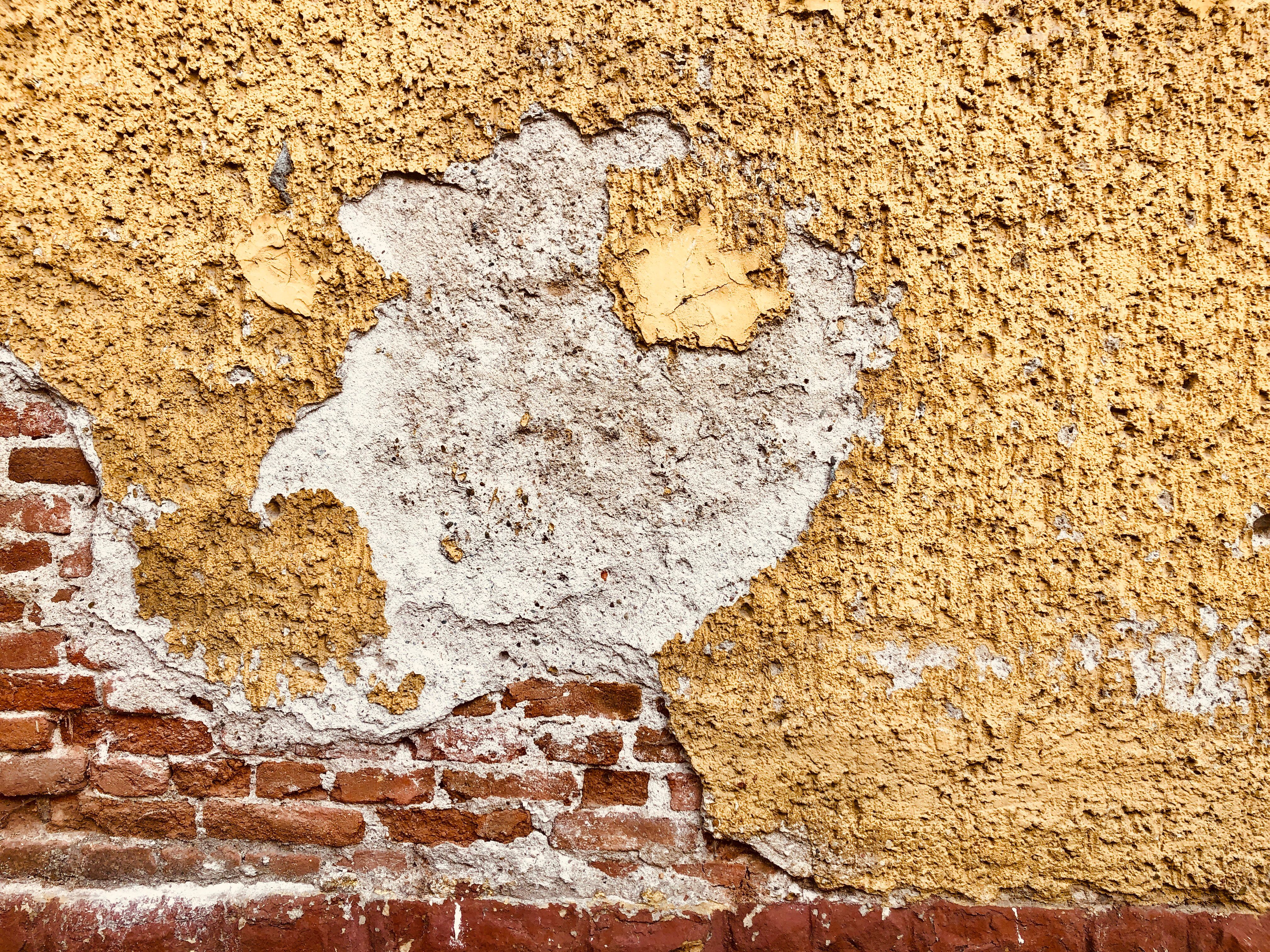 Close-up of a weathered wall showing exposed bricks and peeling plaster with a yellow hue.