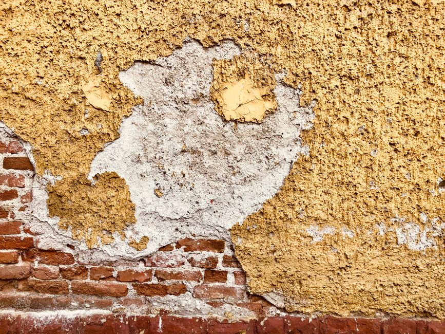 Weathered Mediterranean wall with peeling yellow plaster and exposed brick showing signs of moisture damage and salt mig