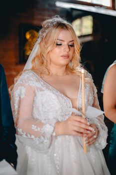 A beautiful bride in an ornate gown holds a candle during a wedding ceremony indoors.