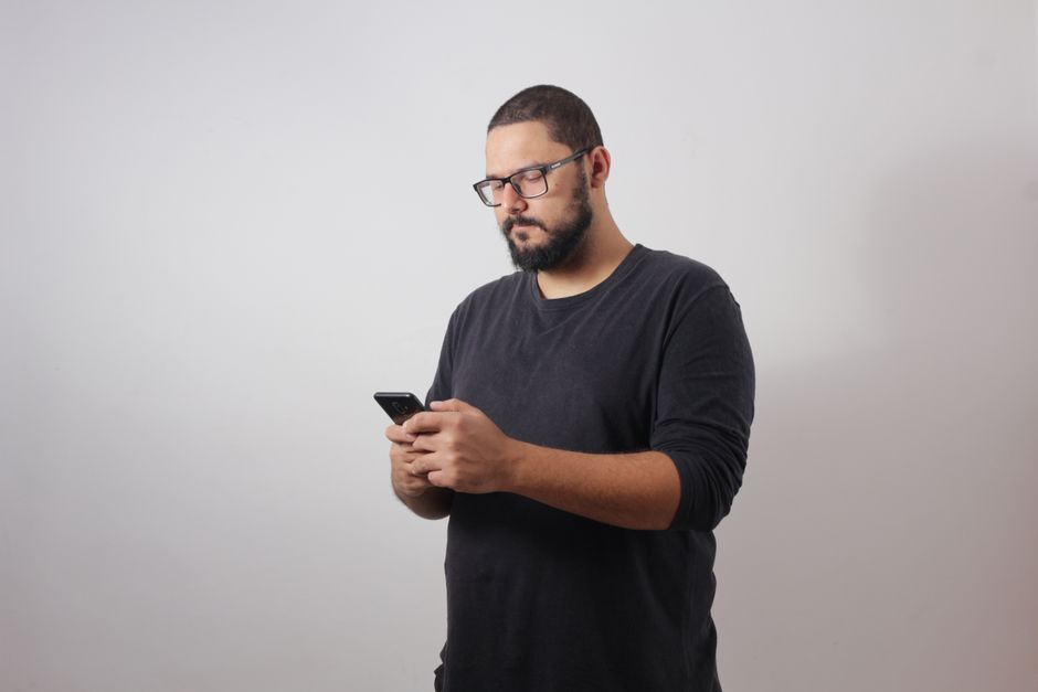 Homem barbudo com óculos usando smartphone em ambiente interno contra fundo neutro.