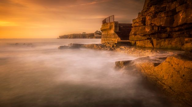 Calm seascape with rocky coast and stairs in golden hour light, creating a peaceful ambiance.