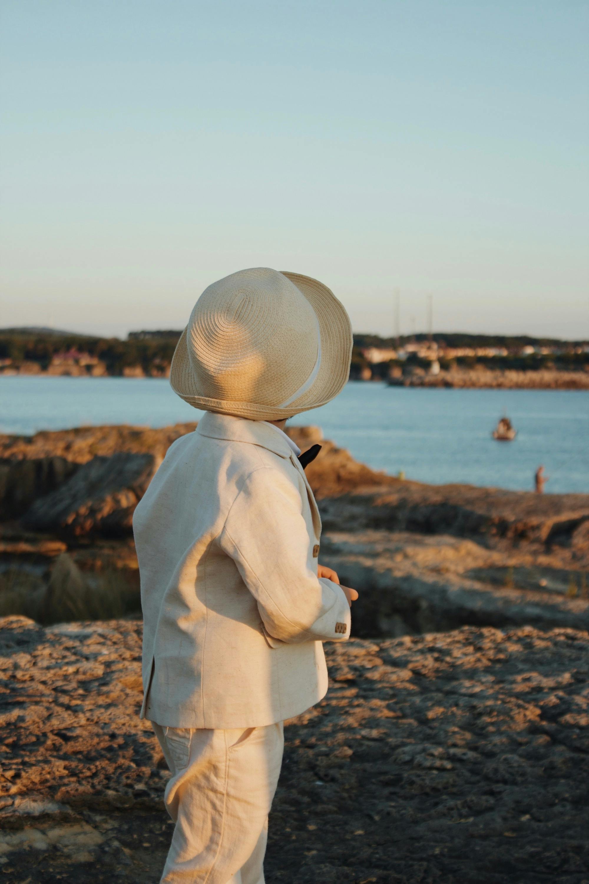Young child in a beige suit and hat gazing at a tranquil lake from rocky land during a sunny day.
