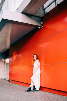 A young woman in a white dress and black boots smiling against a vibrant orange wall.